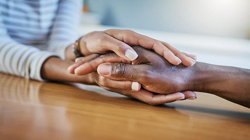 Two people are holding hands on a wooden table while talking in a well-lit space.
