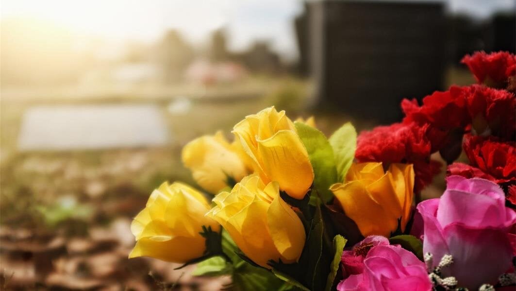 Beautiful yellow, red, and pink flowers rest peacefully at a cemetery as the sun sets behind.