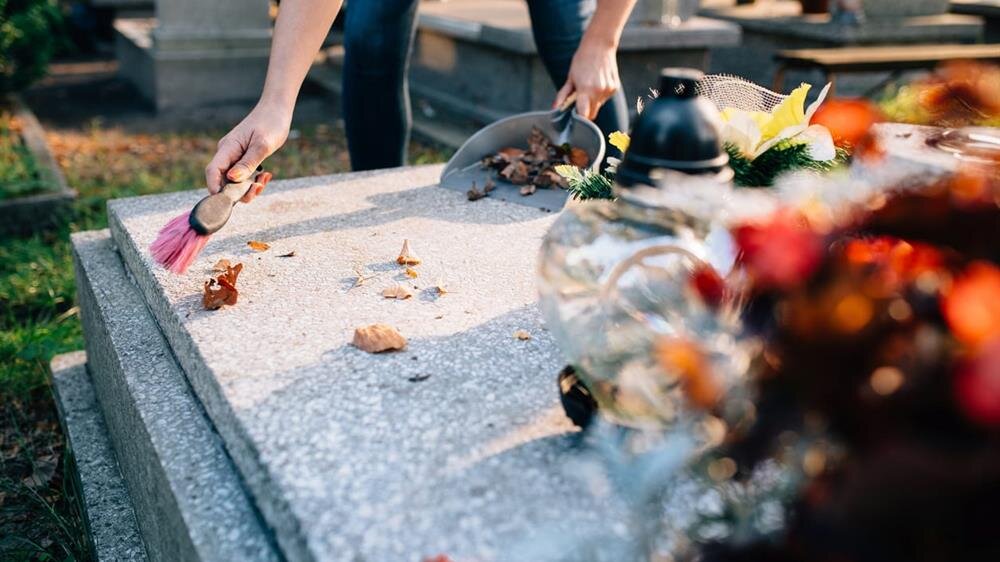 A person gently removes fallen leaves from a grave in a tranquil cemetery setting in fall.
