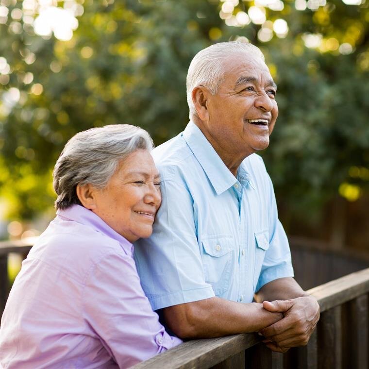 Elderly couple stands close together on a bridge, smiling joyfully while enjoying nature.