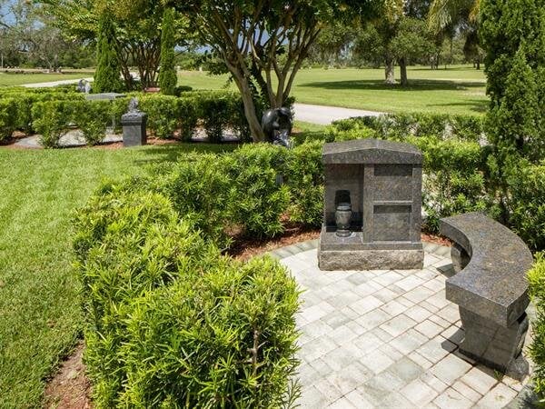 A tranquil garden features a stone bench and memorial among vibrant green shrubs on a sunny day.