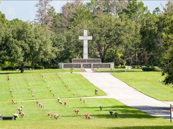 Visitors pay respects in a peaceful memorial park adorned with flowers and a prominent cross.