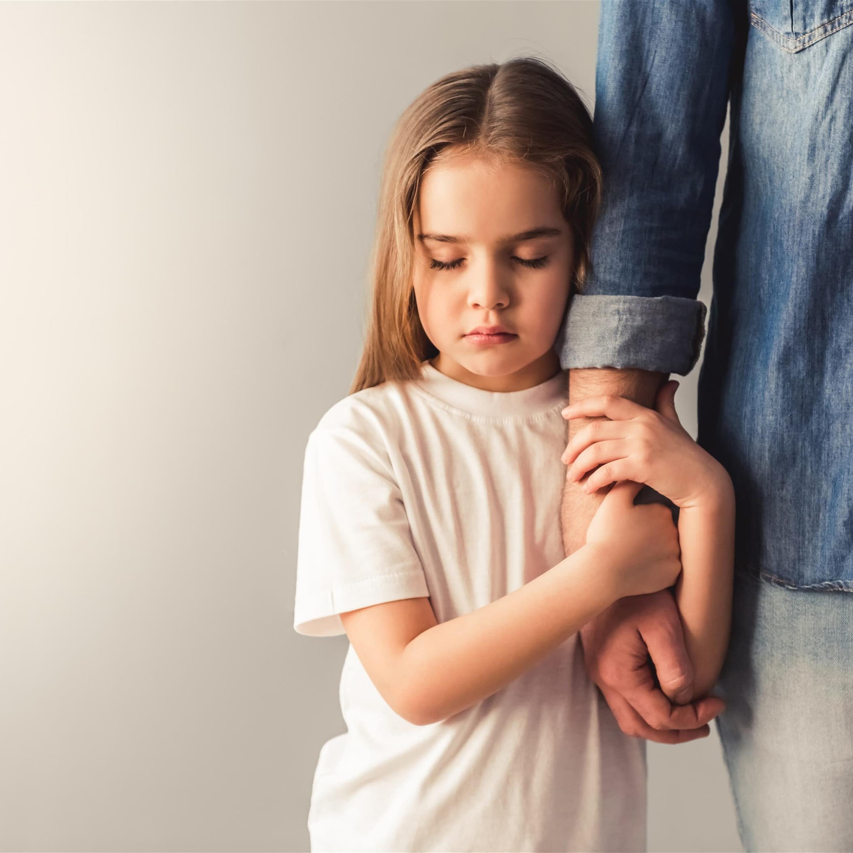 A young child in a white shirt holds a parent's arm, looking down for comfort.