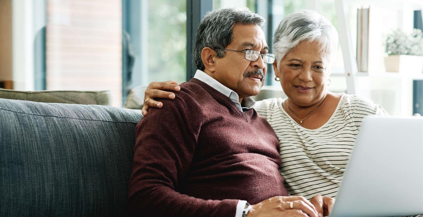 Elderly couple smiles together on the couch while looking at a laptop.