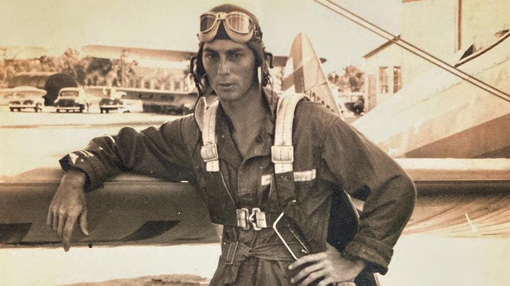 A confident aviator stands beside a classic aircraft, showcasing vintage flying gear and goggles.