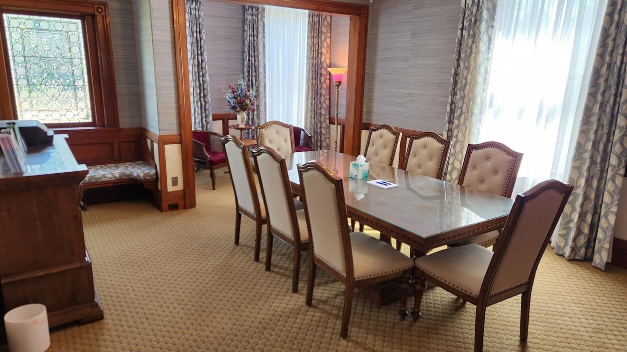 A large dining table surrounded by chairs sits in a room with windows and soft light.