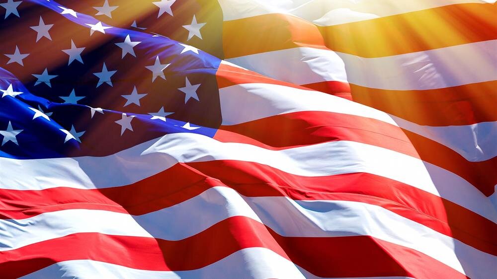 Stars and stripes of the American flag flutter in the wind, illuminated by bright sunlight.