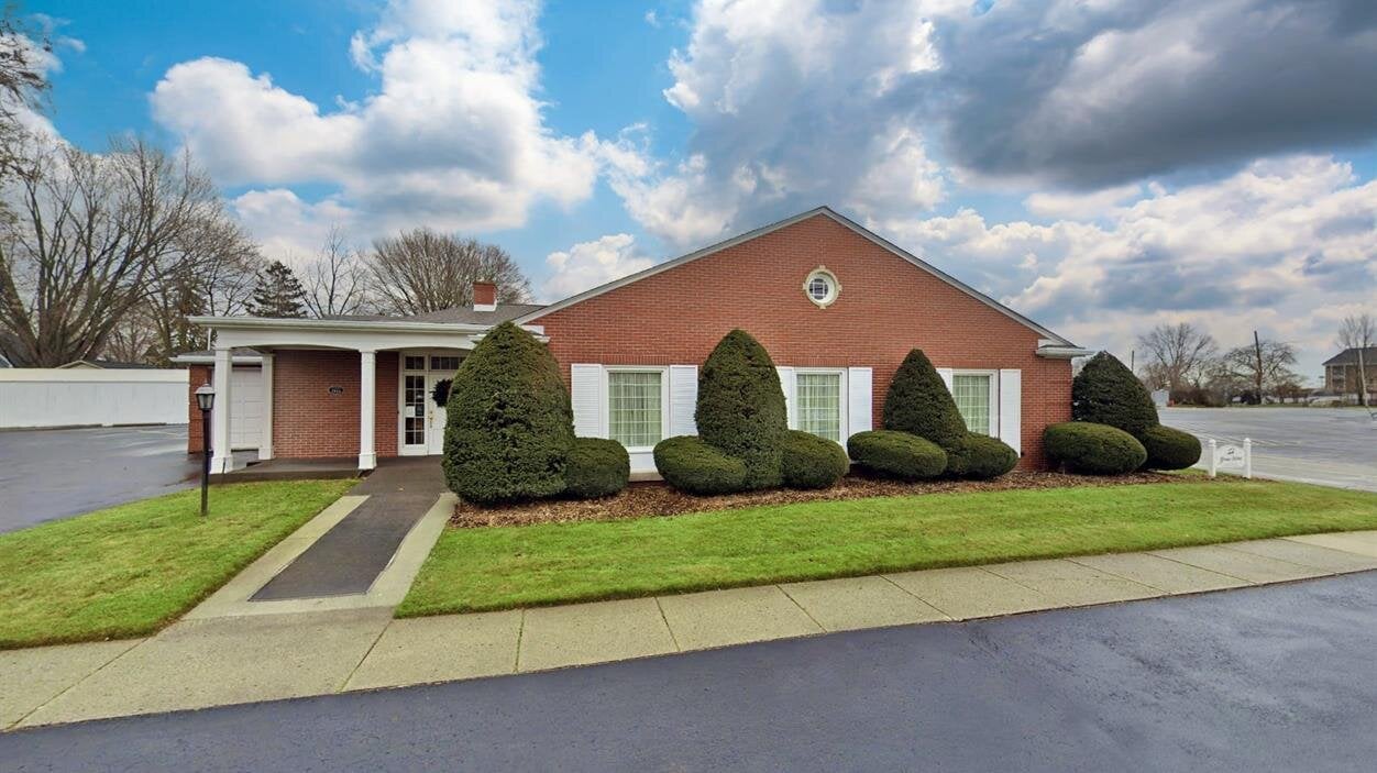 A building sits next to a sidewalk with green grass and bushes under a cloudy sky.
