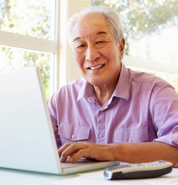 An elderly man sits at a table with a laptop, smiling while wearing headphones during the day.