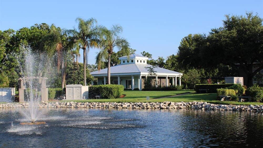 A serene lakeside scene features a lovely house framed by palm trees under clear blue skies.