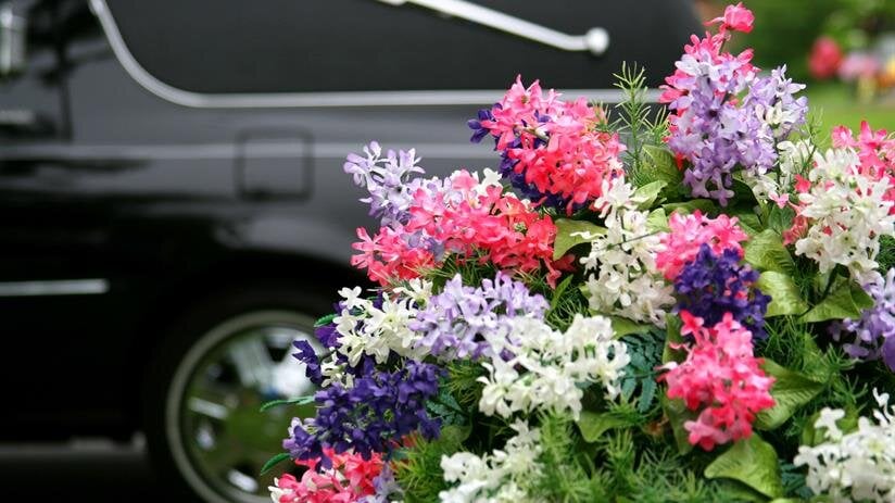Bright flowers are arranged nearby a hearse at a funeral service in a city.