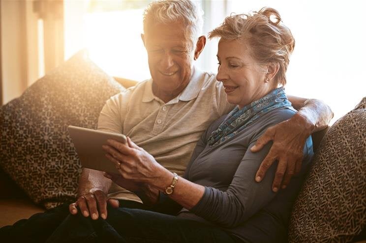A couple sits on a couch together, looking at a tablet and enjoying each other's company.