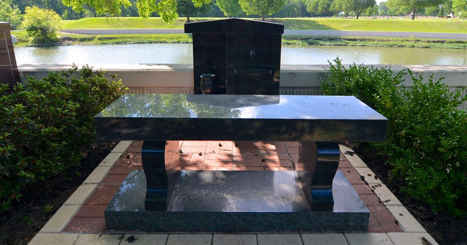 A black granite memorial sits by a peaceful waterway surrounded by trees and green grass.
