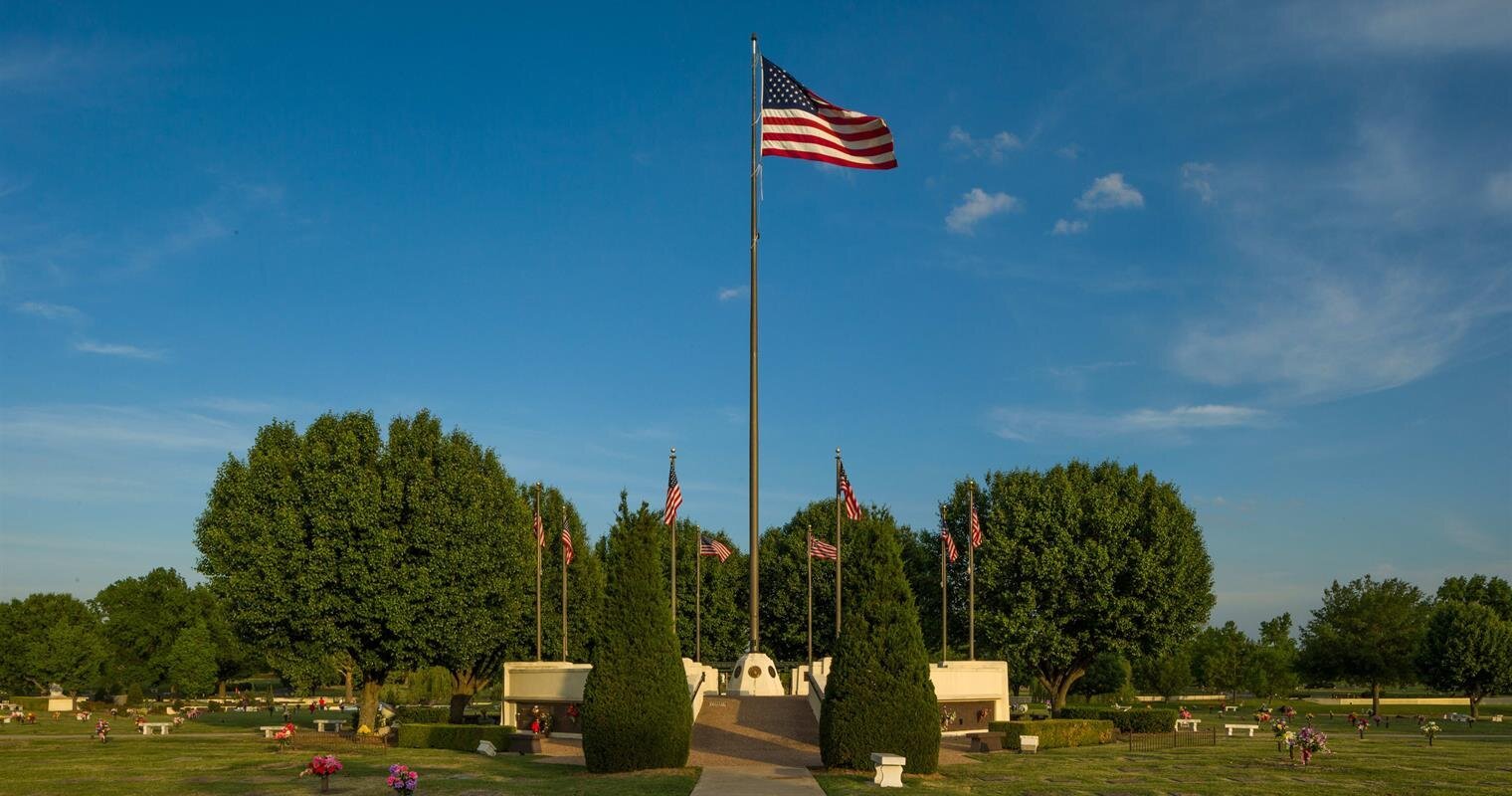 A tall flag stands center stage in a peaceful cemetery, surrounded by trees and a vibrant sky.