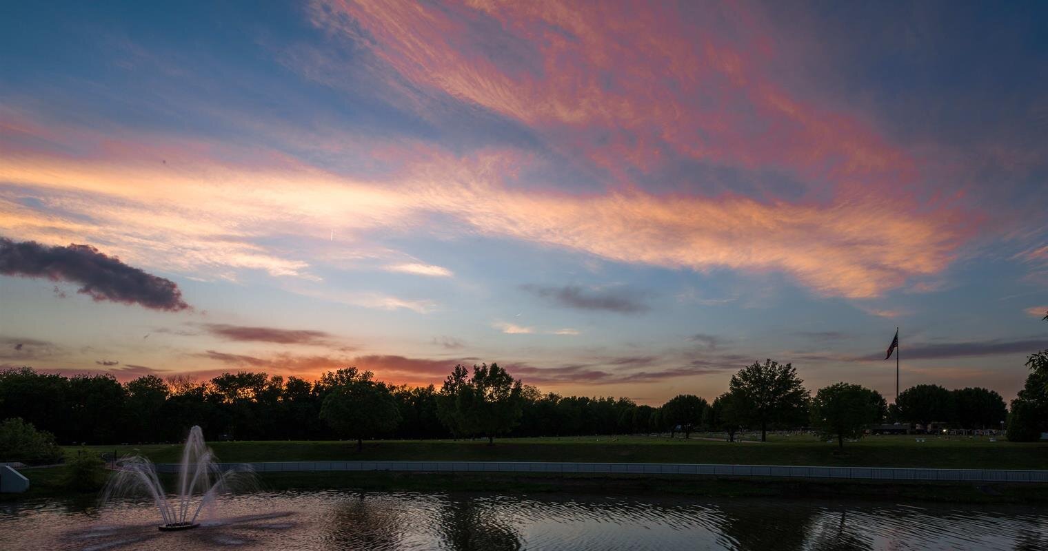 Vibrant clouds stretch across the twilight sky above a tranquil park pond with trees and a fountain.