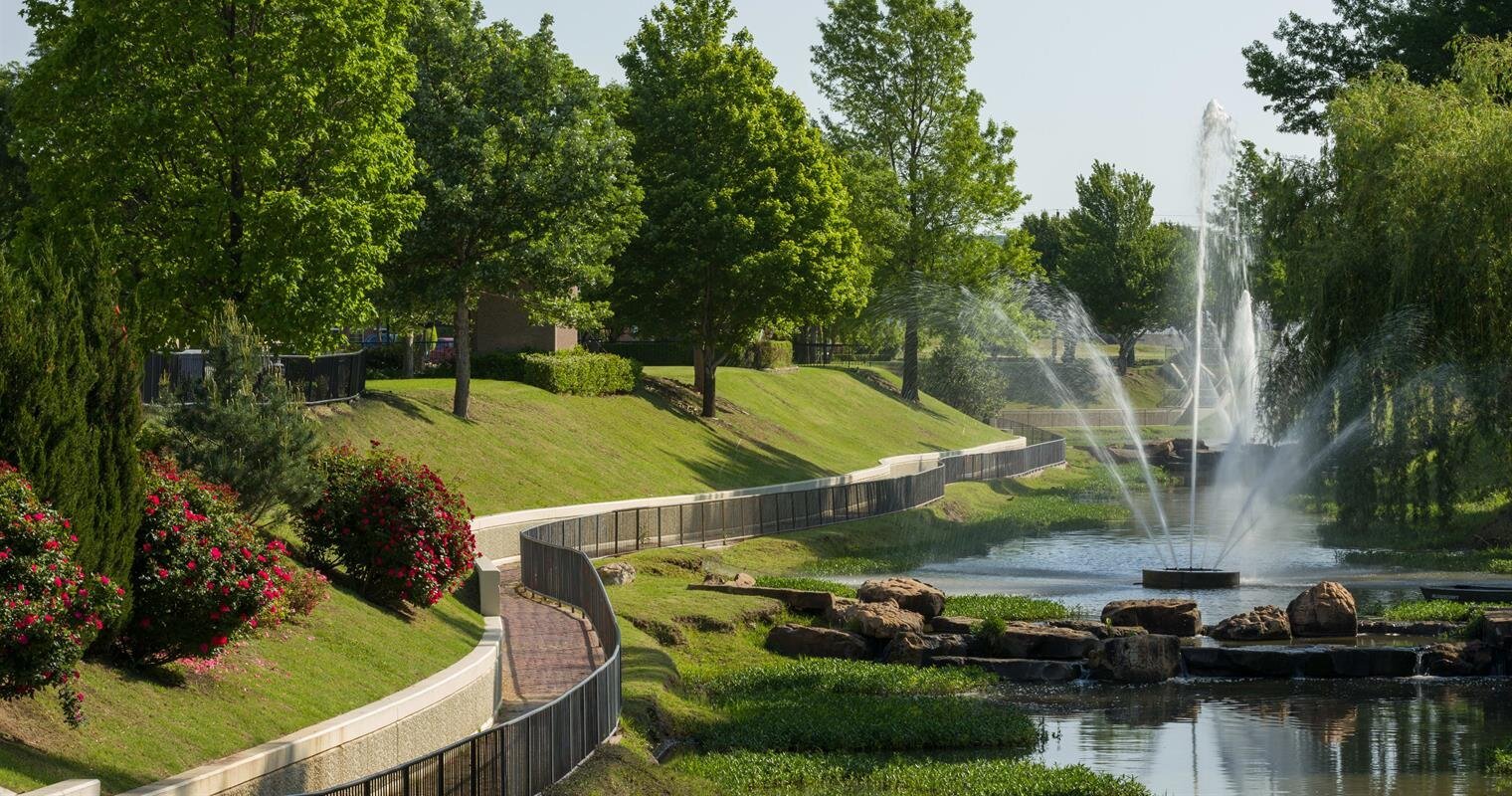 Visitors enjoy a peaceful day by a fountain in a well-maintained park with lush trees.