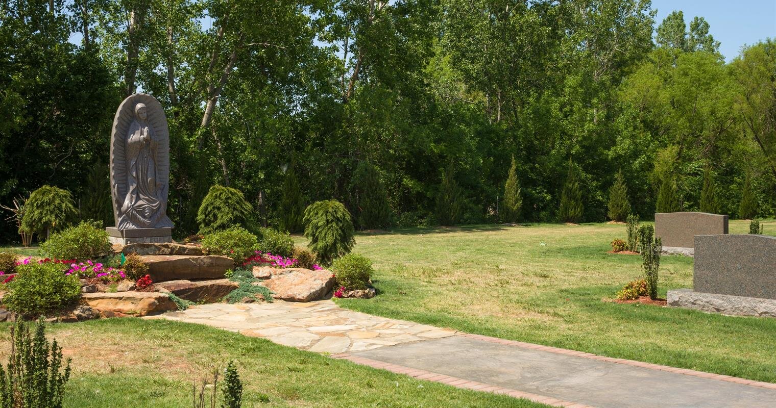 A tranquil park setting showcases a stone statue surrounded by vibrant flowers and green trees.