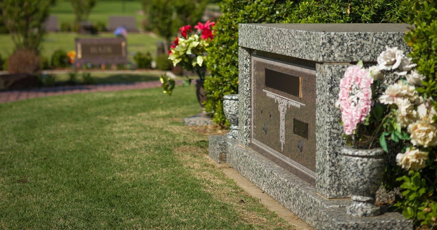 Flowers adorn a grave marker in a peaceful cemetery surrounded by greenery.