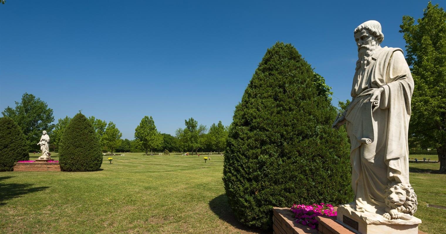 Statues stand in a lush park under a bright blue sky with trimmed hedges and blooming flowers.