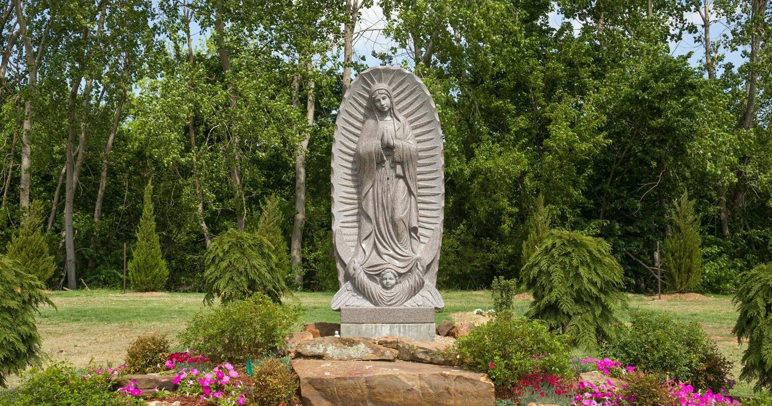 A tall statue of a religious figure stands amid vibrant flowers and lush greenery.