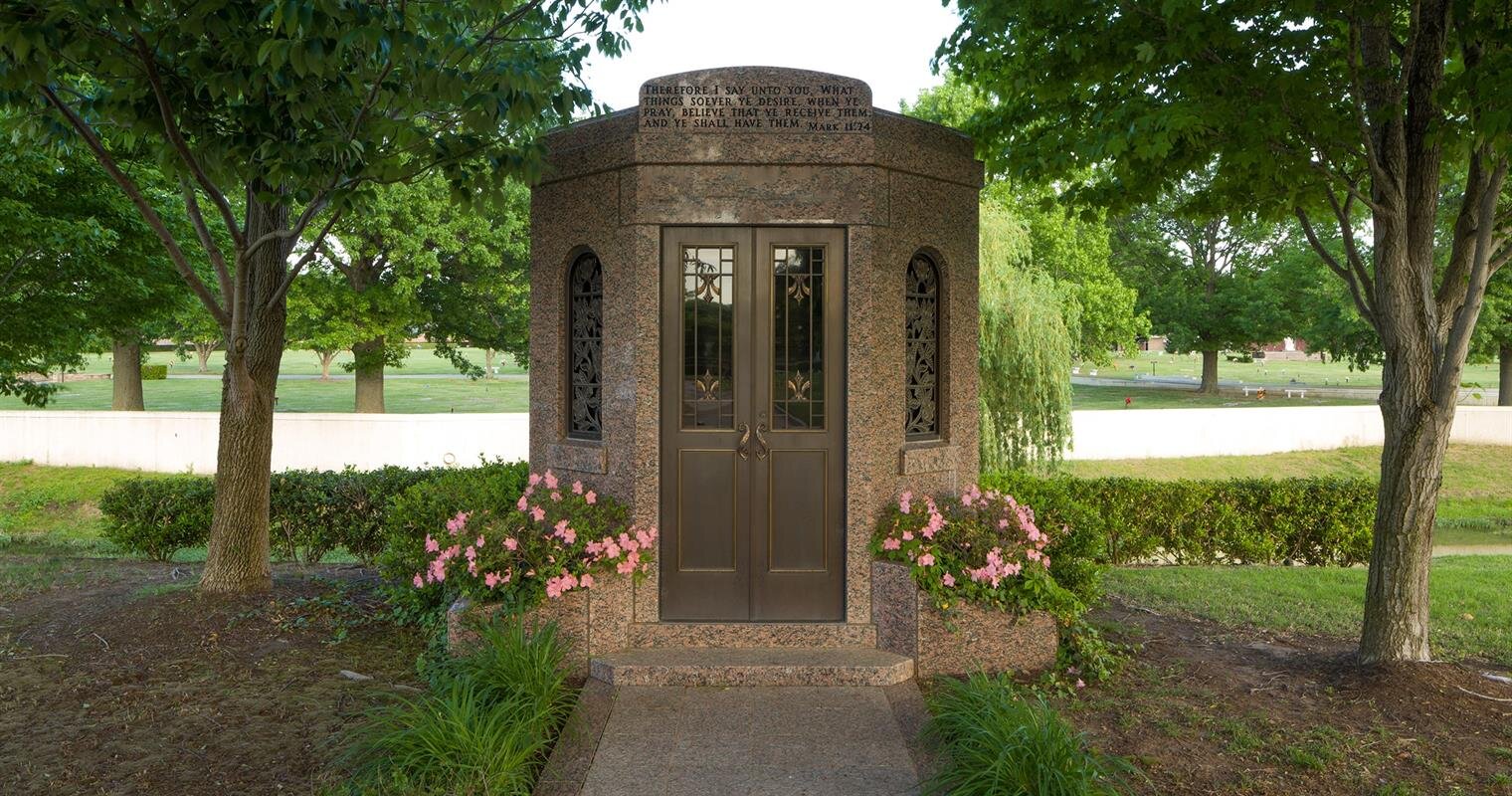 Stone building with double doors stands among vibrant flowers and greenery in a peaceful park.