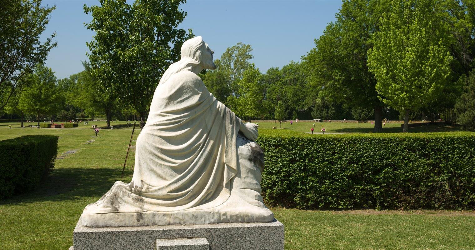 A serene statue sits thoughtfully in a lush park surrounded by greenery and sunlight.