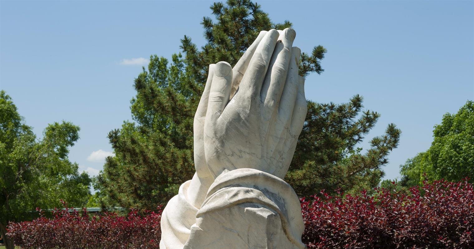 A prominent sculpture of praying hands stands in a tranquil garden, framed by lush trees and bushes.