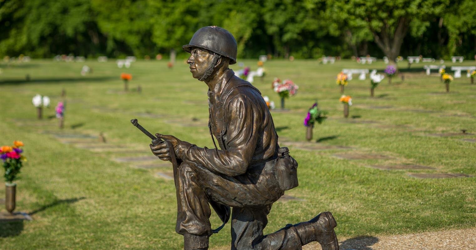 A soldier statue kneels with a radio in a serene cemetery filled with colorful flowers.