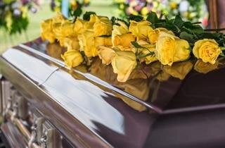 Bright yellow roses lie on a polished casket at a memorial service held in a peaceful location.