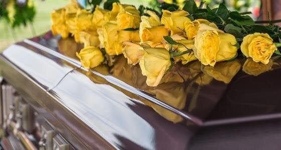 Bright yellow roses lie on a polished casket at a memorial service held in a peaceful location.