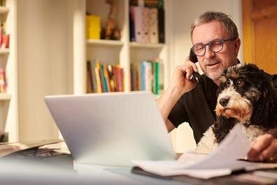A man is working on a laptop, talking on the phone, and holding a dog in his hands at home.