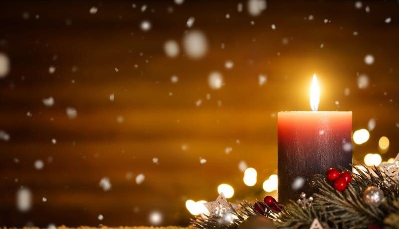 A dark candle shines brightly among holiday decorations as snow gently falls outside.