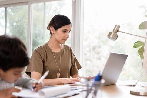 A woman types on her laptop while a child writes beside her at a table in a home office.