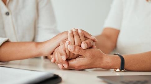 Two individuals are holding hands in a comforting manner during a conversation at work.