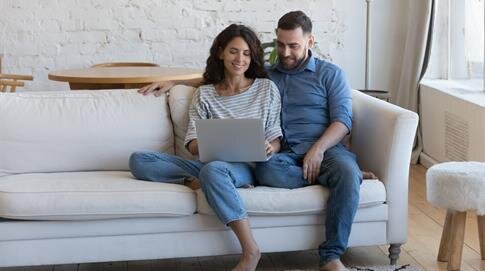 A couple shares smiles and laughter on a sofa while using a laptop in their cozy living room.