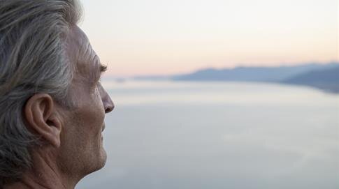 An elderly man admires the tranquil sunset over the calm water, lost in thought.