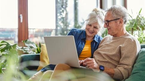 A couple sits closely together on a couch using a laptop in their sunny living room.