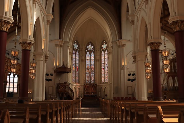 Gothic church interior with tall arches, warm lighting, and stained-glass windows illuminating empty wooden pews.