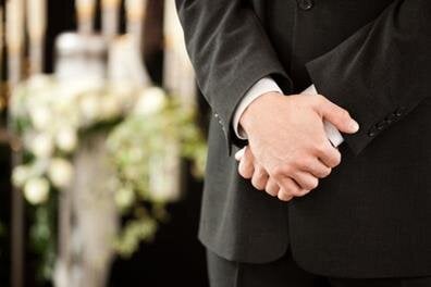A man is standing with hands held together as people gather for a serious occasion.
