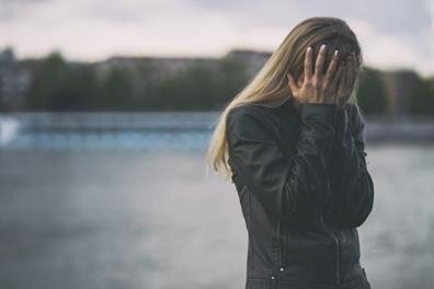 A woman stands near water with her hands on her head while looking down on a cloudy day.