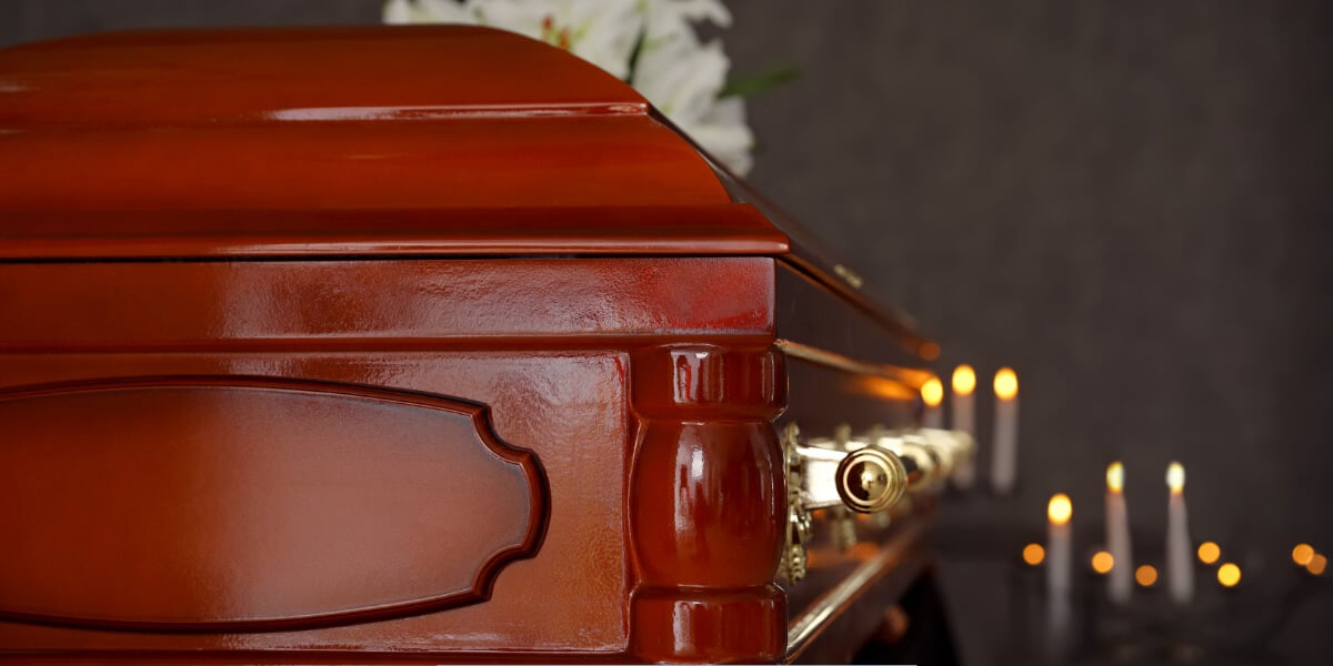 Close-up of a polished wooden casket with brass handles and lit candles in the background during a funeral service.