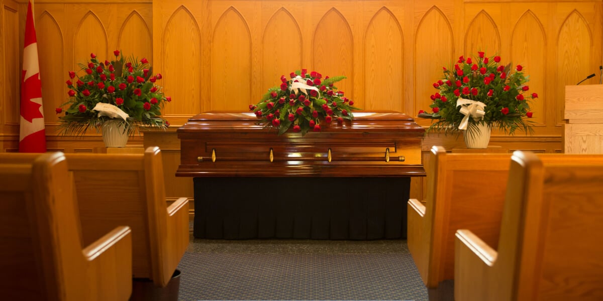 Funeral chapel interior with wooden pews, a polished casket adorned with red rose arrangements, and a Canadian flag displayed beside the altar.