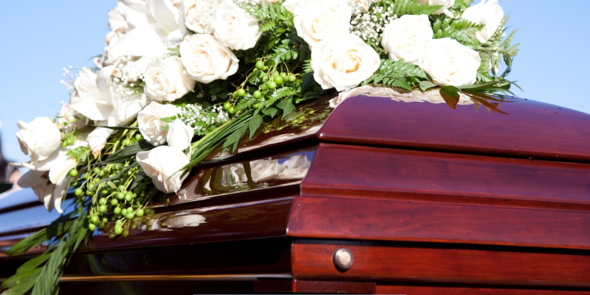 Polished wooden casket topped with a white rose and greenery floral arrangement during an outdoor funeral service under a clear blue sky.