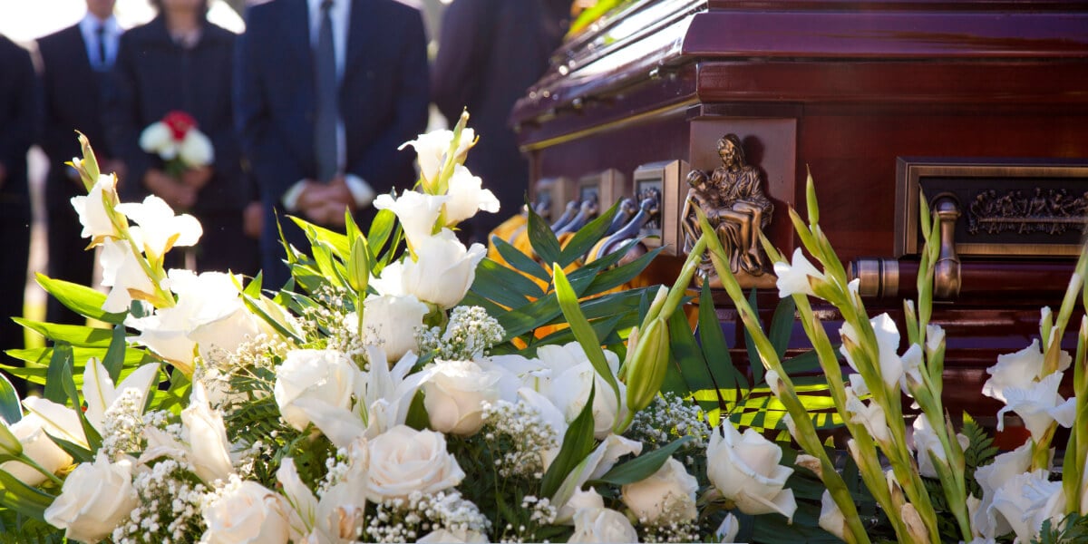 Wooden casket adorned with white roses and lilies at an outdoor funeral service, with mourners standing respectfully in the background.