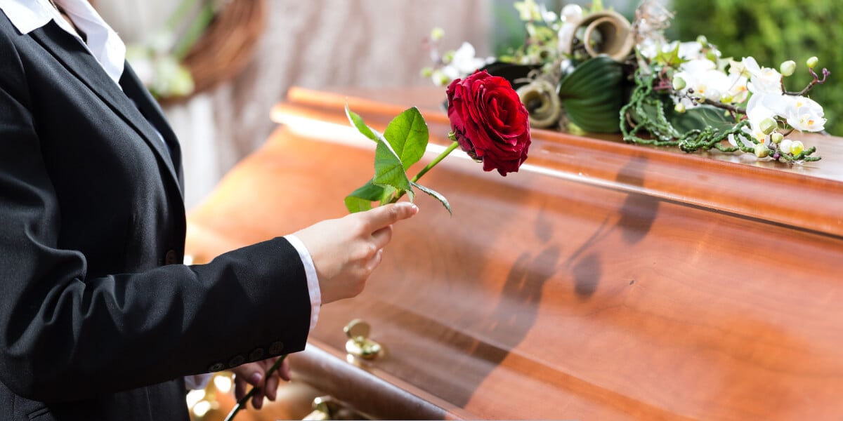 Person in black suit placing a red rose on a polished wooden casket during a funeral service.