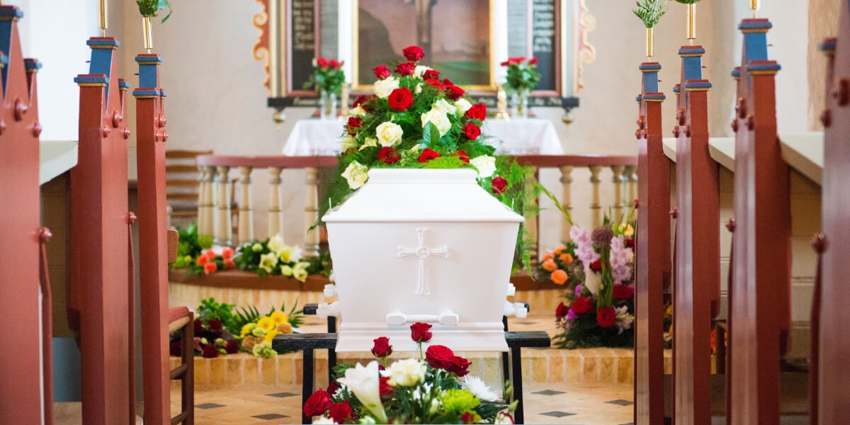 White casket with a cross displayed at the front of a church, surrounded by red and white floral arrangements during a funeral service.