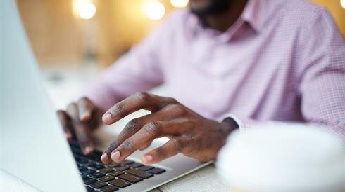 A person types on a laptop while sitting at a cafe table with a drink nearby.
