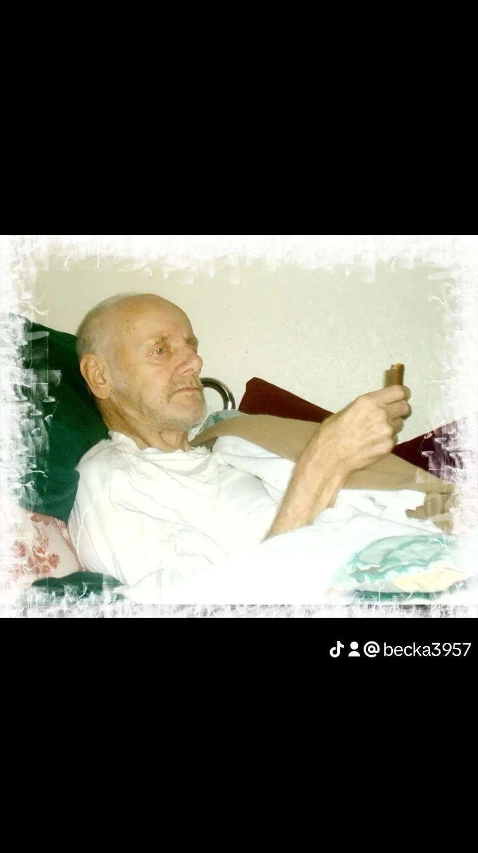 An elderly man lies on a bed, comfortably dressed, calmly holding a small item and resting.