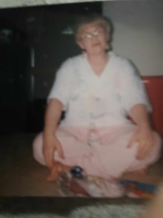 An elderly woman sits on the floor, sewing a patchwork quilt with vibrant fabric pieces around her.