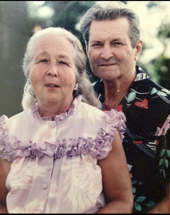 A happy elderly couple smiles warmly at the camera, surrounded by greenery and blossoms.